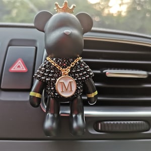 May include: Black bear car air freshener with a gold crown, a gold chain, and a gold pendant with the letter "M" on it. The air freshener is hanging from the air vent of a car.