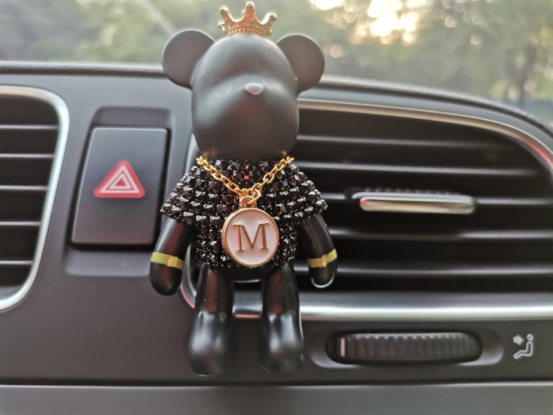 May include: Black bear car air freshener with a gold crown, a gold chain, and a gold pendant with the letter "M" on it. The air freshener is hanging from the air vent of a car.