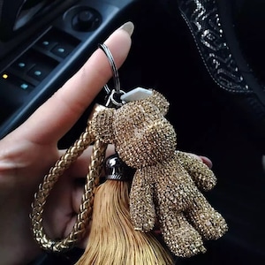 May include: A gold-colored teddy bear keychain with a braided strap and tassel. The bear is covered in sparkling embellishments. The keychain is held up against a dark background.