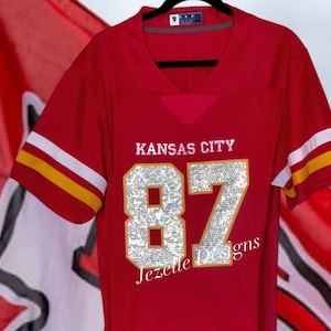 May include: Red Kansas City jersey with the number 87 in silver glitter and gold trim. The text "Jezelle Designs" is below the number. The jersey has white and gold stripes on the sleeves. The text "#custommade" is at the bottom of the image.