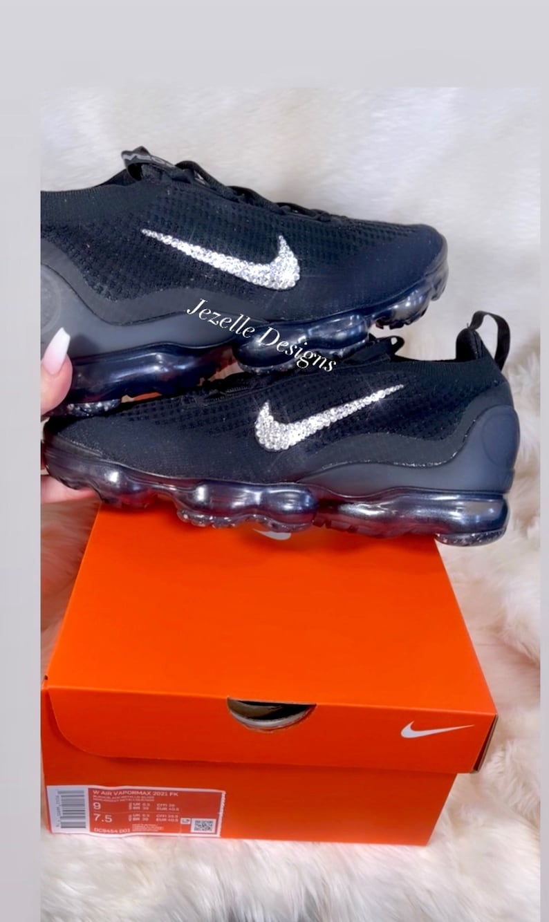 May include: Black Nike Vapormax trainers with a silver rhinestone swoosh logo. The shoes are in a Nike shoebox with a white Nike swoosh logo on the top.