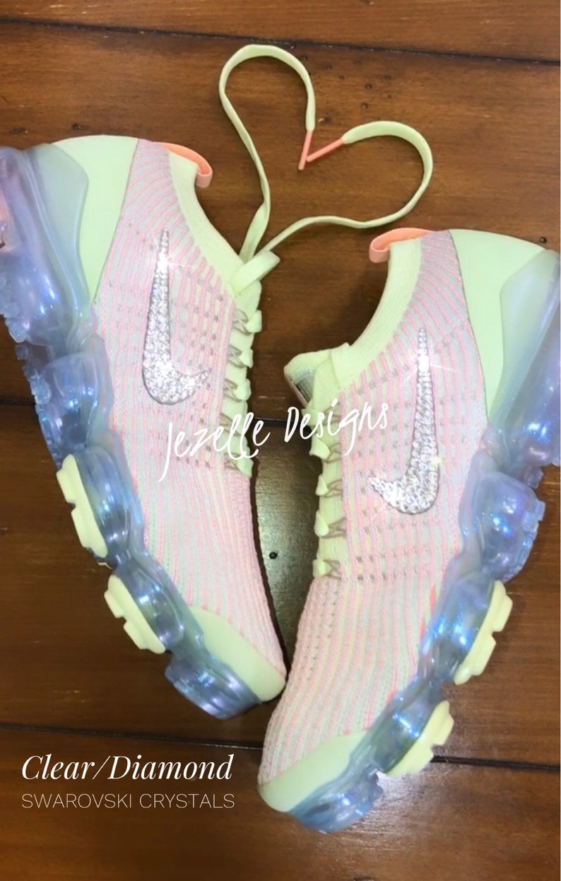 Puede incluir: Un par de zapatillas Nike de color verde claro y rosa con suelas transparentes. El swoosh de Nike est&aacute; decorado con cristales transparentes en forma de diamante. Los cordones est&aacute;n atados en forma de coraz&oacute;n. El texto "Jezelle Designs" es visible en las zapatillas. El texto "Clear/Diamond SWAROVSKI CRYSTALS" es visible debajo de las zapatillas.