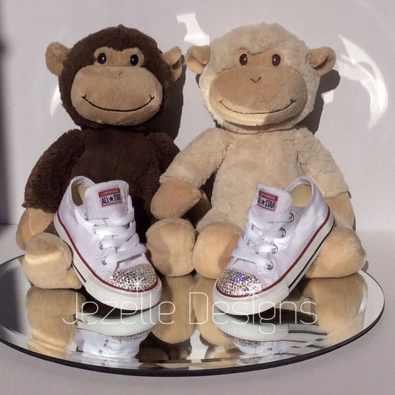 Peut inclure: Deux paires de baskets Converse blanches pour b&eacute;b&eacute; avec des strass sur les orteils. Les baskets sont pos&eacute;es sur une surface miroir devant deux jouets en peluche en forme de singe.