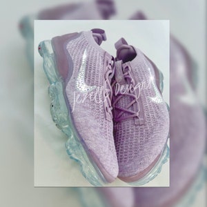 May include: A pair of lavender-coloured trainers with a clear sole and a sparkly Nike swoosh. The trainers are made of a knitted material and have a lace-up closure.