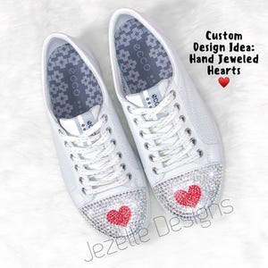 Peut inclure: Une paire de baskets en cuir blanc avec des c&oelig;urs rouges en strass sur les orteils. Les chaussures ont une fermeture &agrave; lacets blanche et une semelle int&eacute;rieure &agrave; motif gris. Le texte "Custom Design Idea: Hand Jeweled Hearts" est &eacute;crit au-dessus des chaussures.