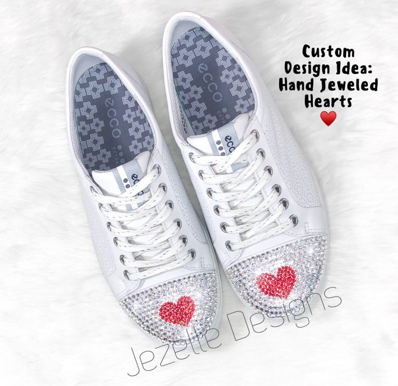 Peut inclure: Une paire de baskets en cuir blanc avec des c&oelig;urs rouges en strass sur les orteils. Les chaussures ont une fermeture &agrave; lacets blanche et une semelle int&eacute;rieure &agrave; motif gris. Le texte "Custom Design Idea: Hand Jeweled Hearts" est &eacute;crit au-dessus des chaussures.