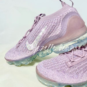 May include: A pair of light purple Nike trainers with a clear sole and a sparkly, crystal-encrusted swoosh logo. The trainers are made of a knit fabric and have a lace-up closure.