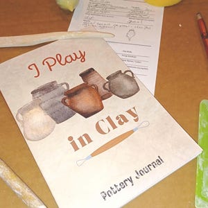 May include: A pottery journal with a light beige cover featuring the text I Play in Clay, illustrations of five ceramic pots, and a clay loop tool. The journal is surrounded by a wooden modeling tool, a red pen, and a green plastic rib tool.