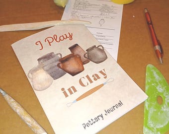 May include: A pottery journal with a light beige cover featuring the text I Play in Clay, illustrations of five ceramic pots, and a clay loop tool. The journal is surrounded by a wooden modeling tool, a red pen, and a green plastic rib tool.