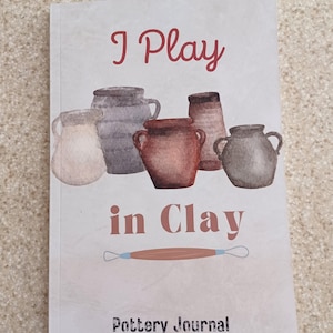 May include: A pottery journal with the text "I Play in Clay" in red and brown. The cover features watercolor illustrations of various clay pots and a pottery tool. The words "Pottery Journal" are printed at the bottom.