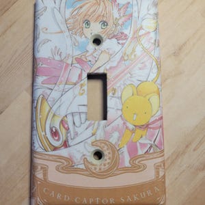 May include: A decorative light switch cover featuring anime artwork. The design includes a character with pink hair and a yellow creature, set against a white and pink background. The text "CARD CAPTOR SAKURA" is at the bottom.