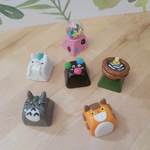 May include: Six colorful, hand-painted keycaps featuring various characters and designs. The keycaps are made of polymer clay and are designed to fit standard mechanical keyboards. The keycaps include a cat, a bird, a Totoro, a unicorn, a monster, and a gumball machine.