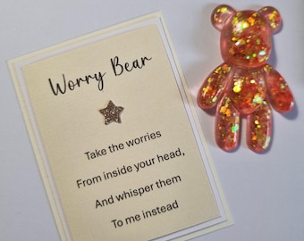 Op de afbeelding: Een helder, met glitter gevuld harsbeerfiguur naast een kaart. De kaart leest "Worry Bear" en bevat tekst die de gebruiker aanmoedigt om zijn zorgen te delen. De beer is gevuld met oranje en gouden glitter.