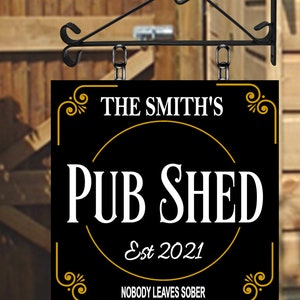 May include: Black rectangular sign with gold trim and text "THE SMITH'S PUB SHED Est 2021 NOBODY LEAVES SOBER." The sign hangs from a black metal bracket. The background is a wooden fence.