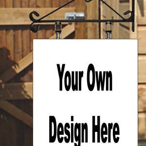 May include: A blank white sign with black text that reads "Your Own Design Here" hanging from a black metal sign bracket. The sign is hanging in front of a wooden fence.