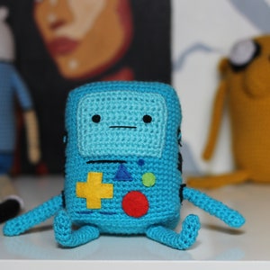 Puede incluir: Un juguete de crochet azul con forma de consola de videojuegos con un signo más amarillo, un círculo rojo y un círculo verde en la parte delantera. El juguete tiene una línea negra para la boca y dos ojos negros.