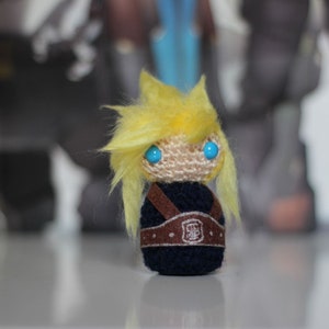 Puede incluir: Un juguete de peluche tejido a crochet de un personaje con cabello amarillo, ojos azules y un atuendo negro y marrón. El juguete lleva un cinturón marrón con una hebilla plateada.