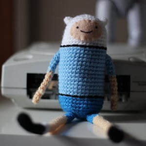 May include: A blue and white crocheted plush toy with a smiling face. The toy is sitting on a white surface.