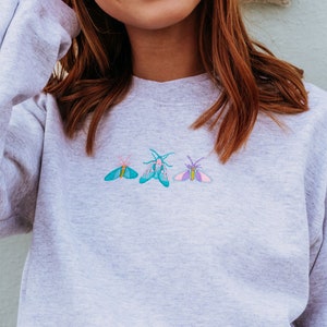K&ouml;nnte beinhalten: Ein graues Sweatshirt mit drei gestickten Motten in verschiedenen Farben. Die Motten sind in einer Reihe angeordnet, wobei die mittlere Motte die gr&ouml;&szlig;te ist. Alle Motten sind nach rechts gerichtet.