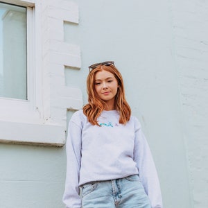 K&ouml;nnte beinhalten: Eine Frau tr&auml;gt ein hellgraues Sweatshirt mit einem bunten Schmetterlingsdesign und hellblaue Jeans-Shorts. Sie steht vor einer wei&szlig;en und hellblauen Wand.