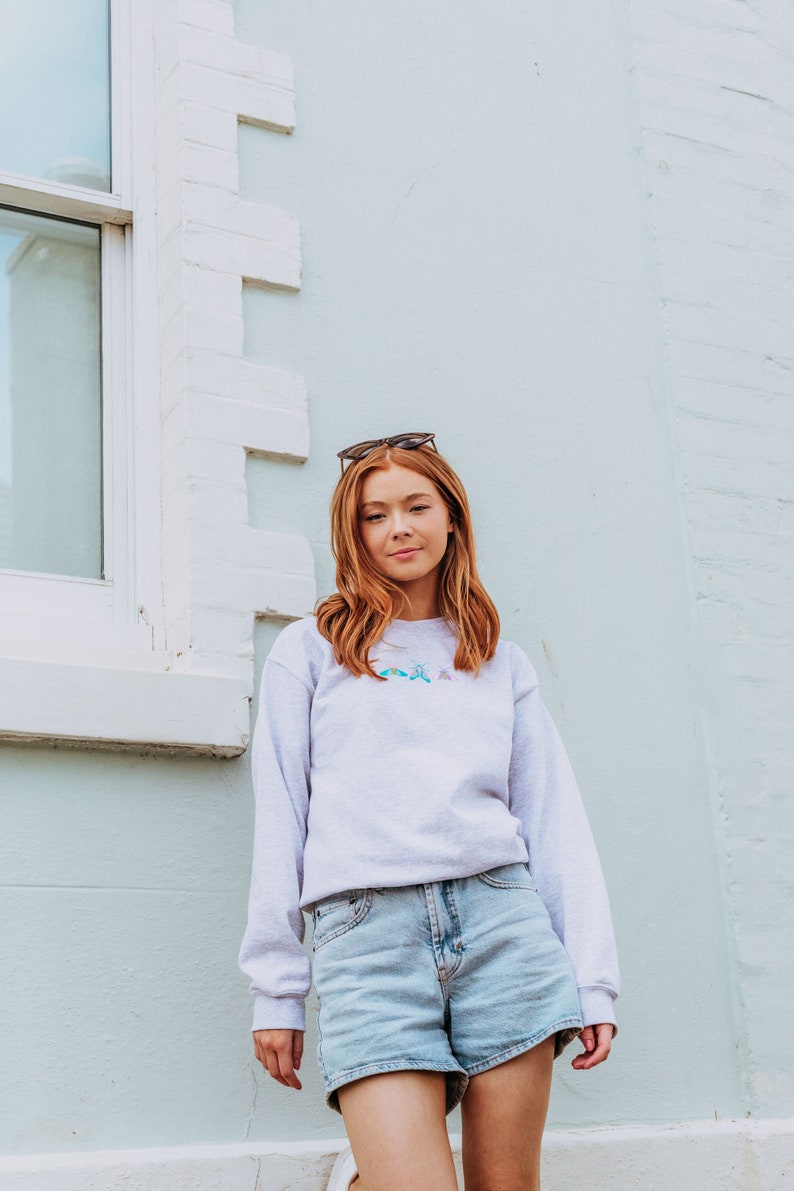 K&ouml;nnte beinhalten: Eine Frau tr&auml;gt ein hellgraues Sweatshirt mit einem bunten Schmetterlingsdesign und hellblaue Jeans-Shorts. Sie steht vor einer wei&szlig;en und hellblauen Wand.