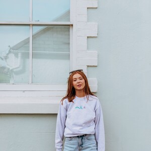 K&ouml;nnte beinhalten: Eine Frau tr&auml;gt ein hellgraues Sweatshirt mit einem bunten Stickmuster und eine hellblaue Jeans-Shorts. Sie steht vor einer wei&szlig;en Backsteinwand mit einem Fenster hinter ihr.
