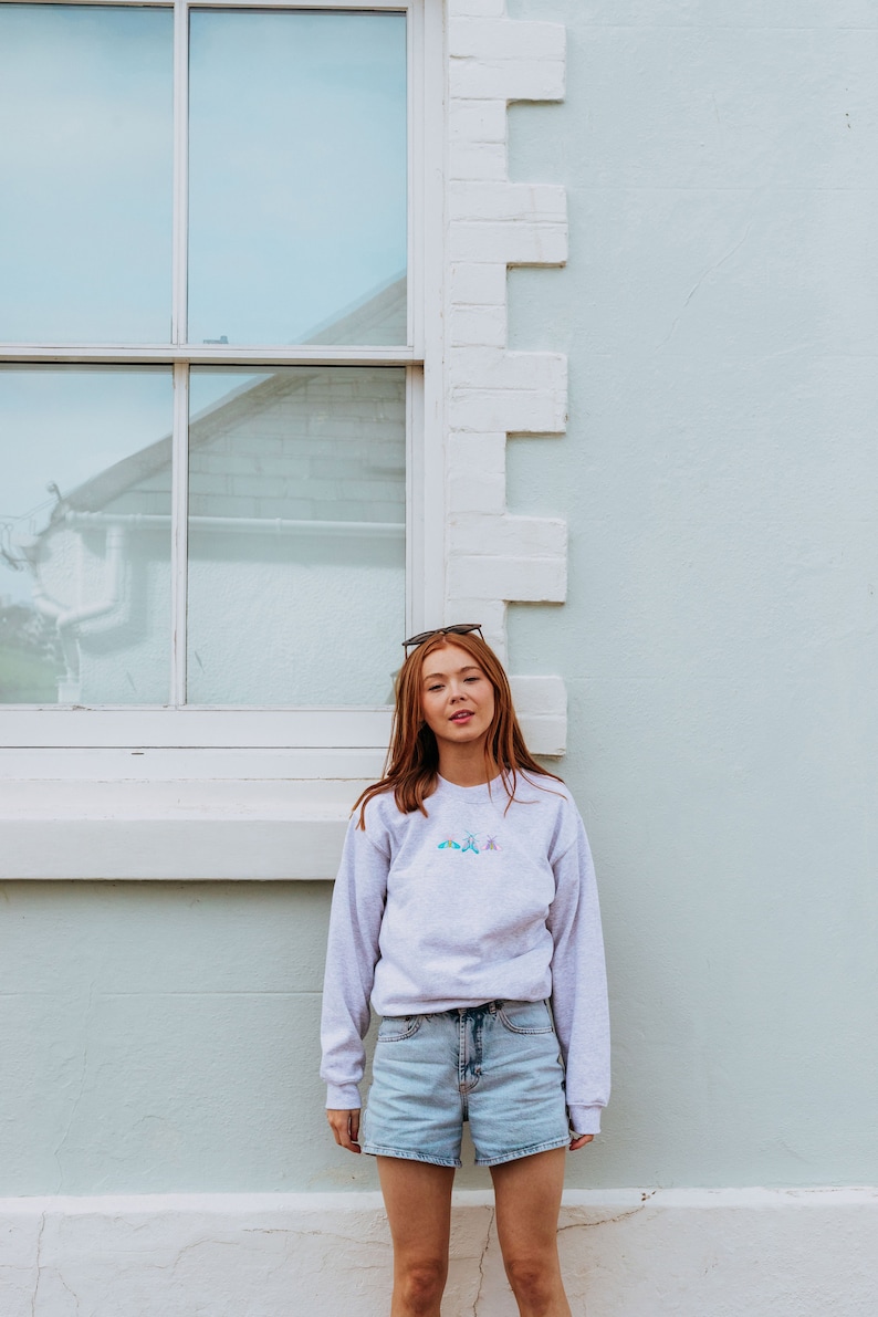 K&ouml;nnte beinhalten: Eine Frau tr&auml;gt ein hellgraues Sweatshirt mit einem bunten Stickmuster und eine hellblaue Jeans-Shorts. Sie steht vor einer wei&szlig;en Backsteinwand mit einem Fenster hinter ihr.