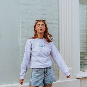 K&ouml;nnte beinhalten: Eine Frau tr&auml;gt ein graues Sweatshirt mit einem kleinen gestickten Design aus drei Schmetterlingen in Blaut&ouml;nen, Rosa und Gr&uuml;n. Sie tr&auml;gt auch hellblaue Jeans-Shorts.
