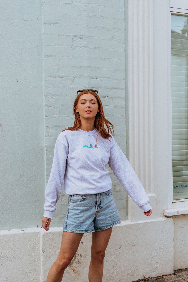 K&ouml;nnte beinhalten: Eine Frau tr&auml;gt ein graues Sweatshirt mit einem kleinen gestickten Design aus drei Schmetterlingen in Blaut&ouml;nen, Rosa und Gr&uuml;n. Sie tr&auml;gt auch hellblaue Jeans-Shorts.