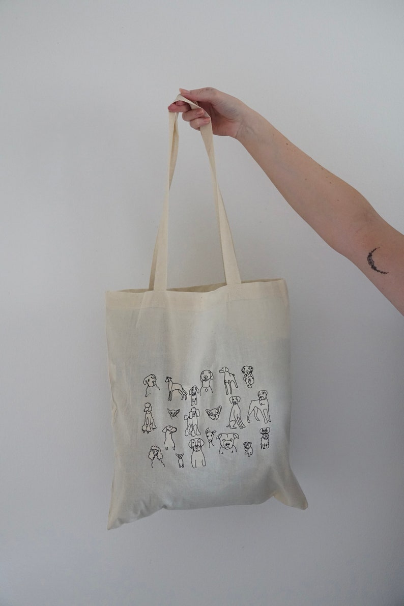 May include: A white canvas tote bag with black line drawings of various dog breeds. The bag has two long straps.