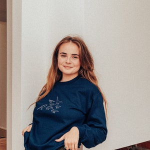 May include: A woman wearing a navy blue sweatshirt with a white embroidered design of dinosaurs. She is wearing gold sparkly pants and is standing in front of a white wall.