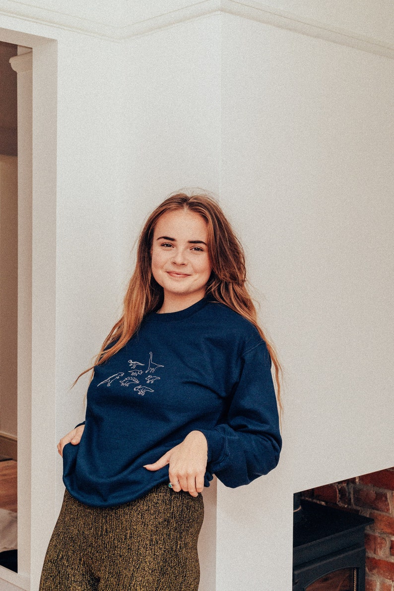 May include: A woman wearing a navy blue sweatshirt with a white embroidered design of dinosaurs. She is wearing gold sparkly pants and is standing in front of a white wall.
