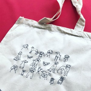 May include: A white canvas tote bag with a black embroidered design of various dog breeds. The bag has two long straps.