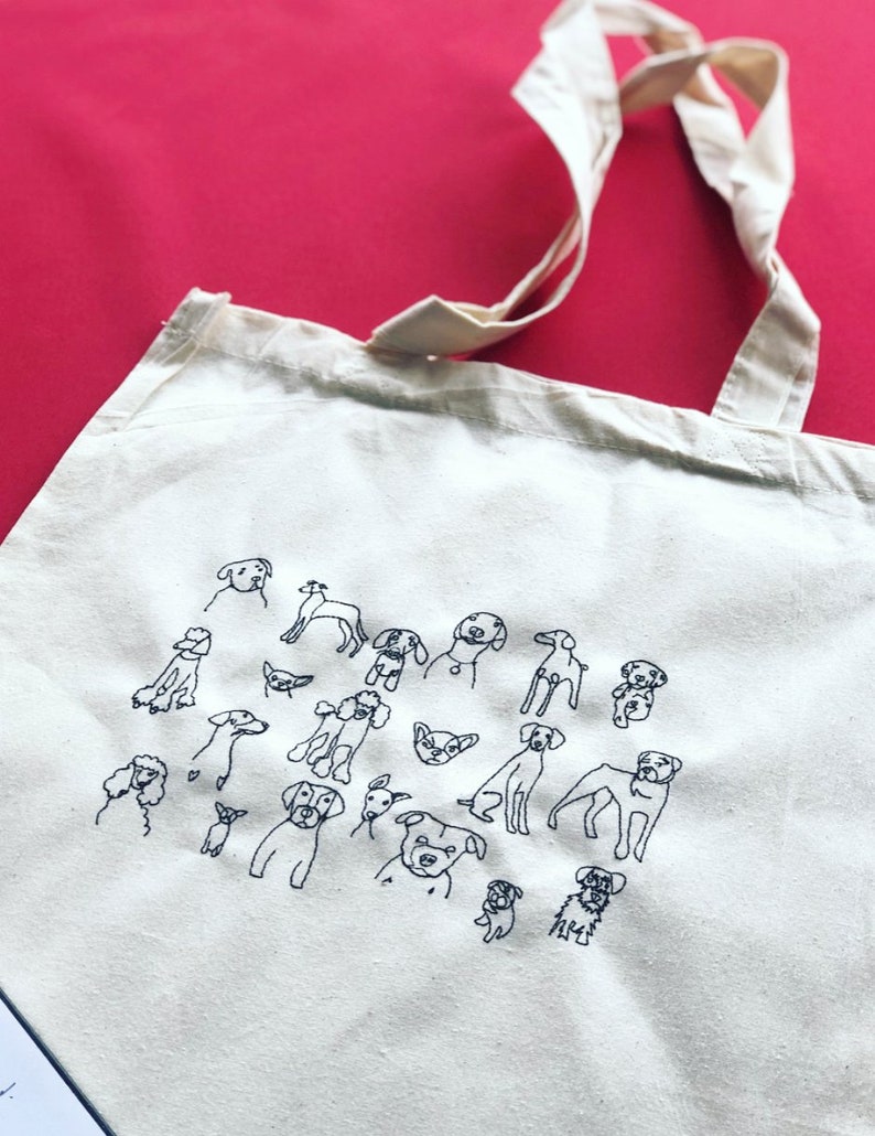 May include: A white canvas tote bag with a black embroidered design of various dog breeds. The bag has two long straps.