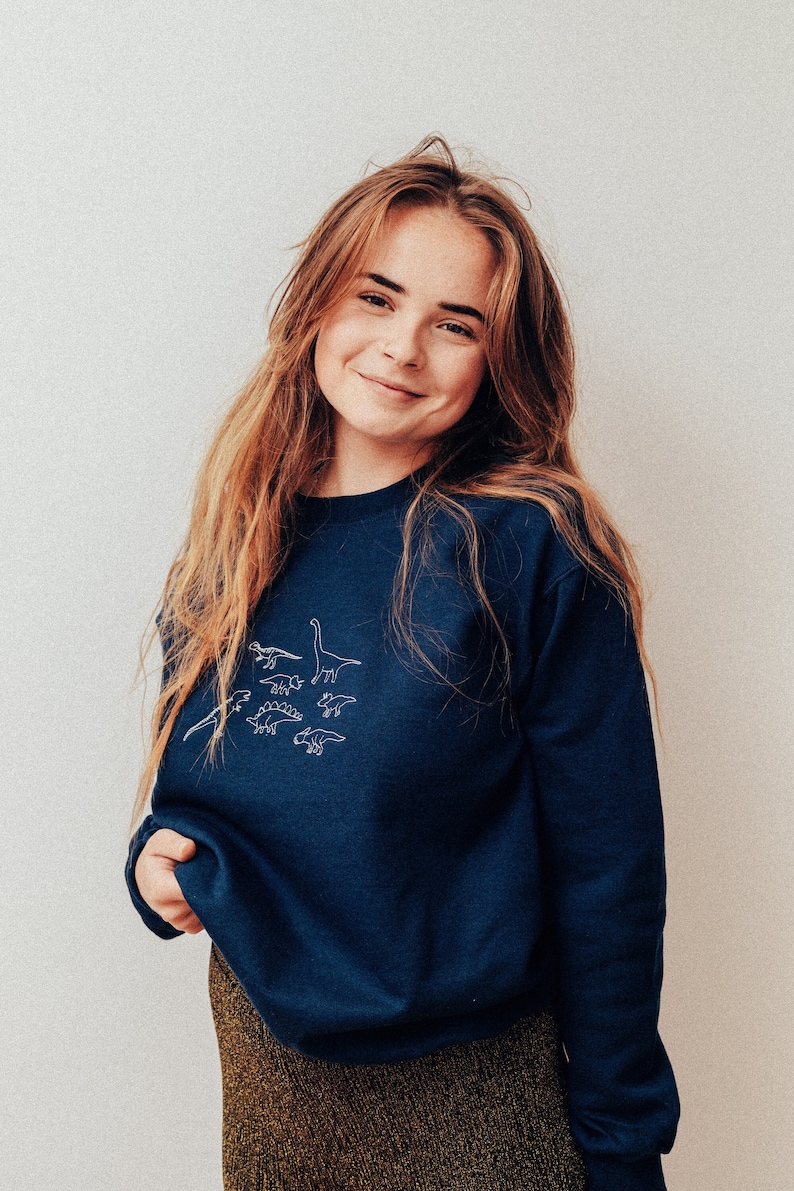 May include: A woman with long brown hair is wearing a navy blue sweatshirt with a white outline of dinosaurs on the front. She is smiling and looking at the camera. She is wearing a gold skirt.