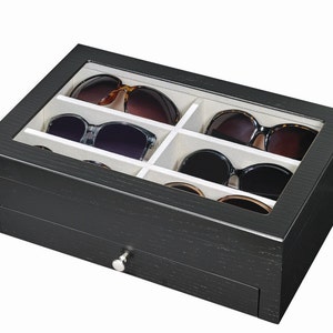 Peut inclure: Un organisateur de lunettes de soleil en bois noir avec un dessus en verre, comprenant six compartiments doublés de tissu blanc cassé. L'organisateur comprend un petit tiroir avec un bouton argenté et contient plusieurs paires de lunettes de soleil.
