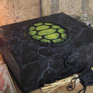 Könnte beinhalten: Eine schwarze, strukturierte Box mit einem grün-gelben kreisförmigen Design auf dem Deckel. Die Box hat einen Metallverschluss und ist von Kerzen, Tarotkarten und einem kleinen schwarzen Beutel umgeben. Die Box wird wahrscheinlich zur Aufbewahrung verwendet.