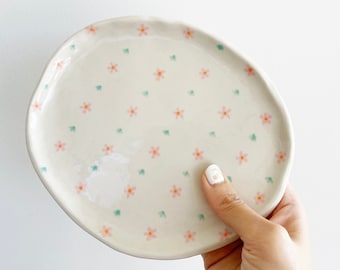 May include: A small, white ceramic plate with a scalloped edge. The plate is decorated with a repeating pattern of pink flowers with green leaves.