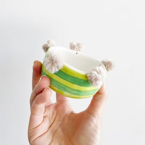May include: A small, green and yellow striped ceramic bowl with a white interior. The bowl has a row of fluffy, light brown pom-poms around the rim.
