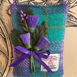May include: A small, rectangular pouch made of teal and purple plaid Harris Tweed fabric. It features a purple cord strap and is adorned with artificial purple thistles, green leaves, and a purple ribbon. A white label with red text is attached.
