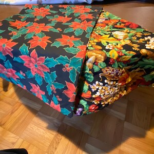 May include: Two tablecloths, one with a red poinsettia pattern and the other with a fall harvest theme, are folded on a wooden table.