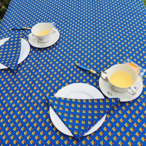 May include: Two place settings on a blue tablecloth with a yellow and red floral pattern. Each setting includes a white plate, a folded napkin, a teacup and saucer, and a spoon.