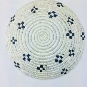 May include: A round woven basket with a white background and black geometric patterns. The basket is made of natural materials and has a circular shape.