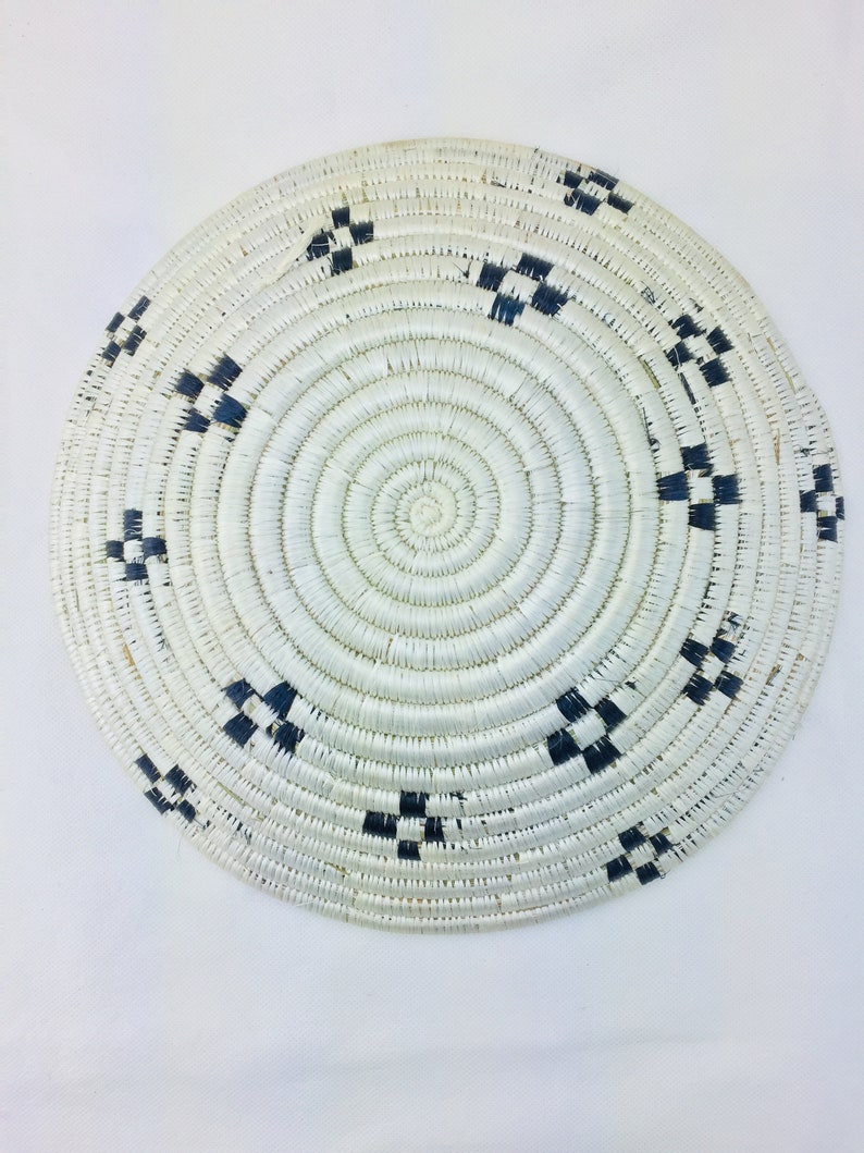 May include: A round woven basket with a white background and black geometric patterns. The basket is made of natural materials and has a circular shape.