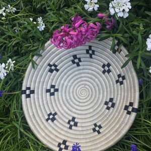 May include: A white woven basket with black trim, featuring a circular design. The basket is surrounded by pink and white flowers.