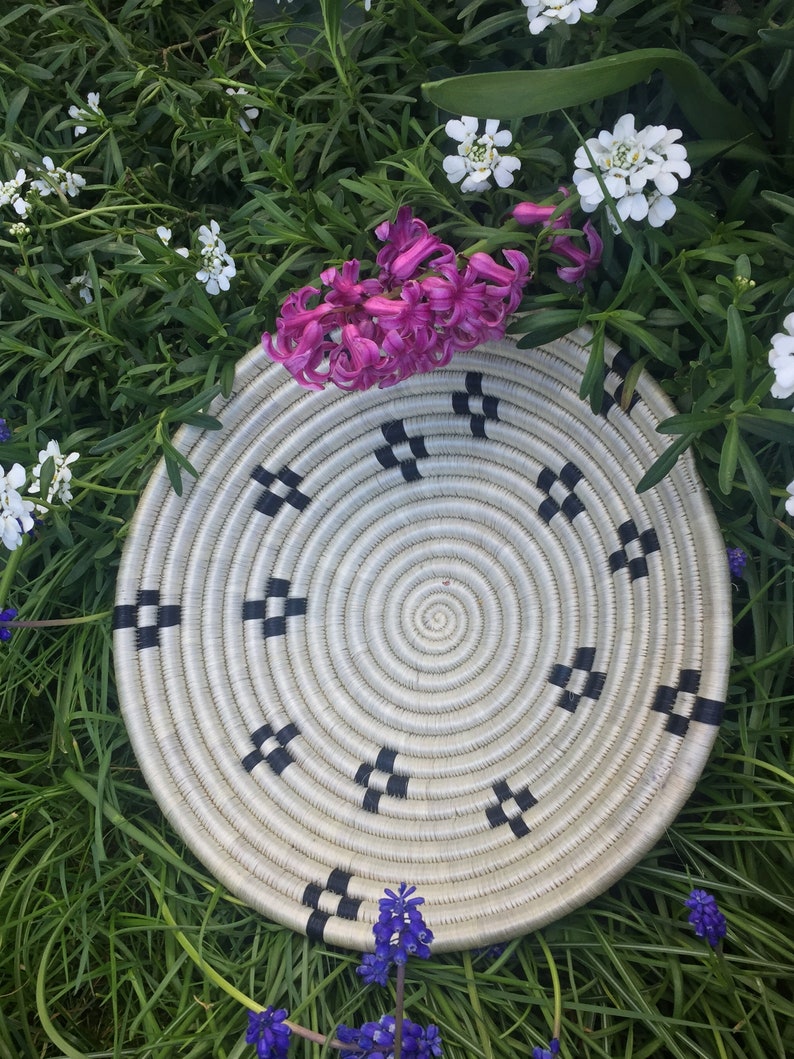 May include: A white woven basket with black trim, featuring a circular design. The basket is surrounded by pink and white flowers.