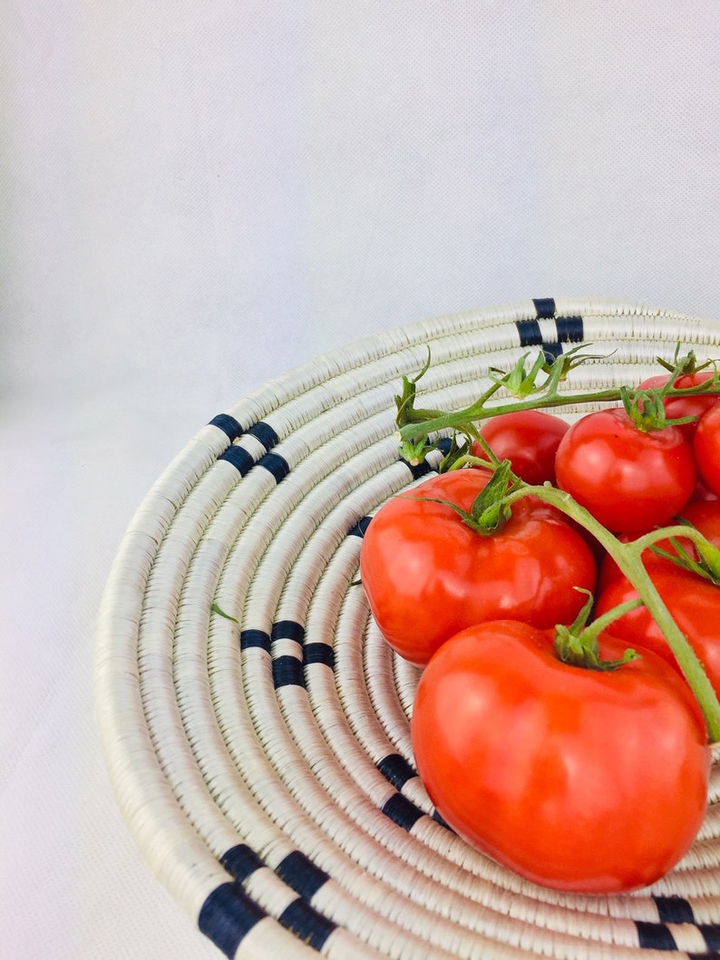 May include: A white woven basket with blue stripes holds a cluster of red ripe tomatoes. The tomatoes are still attached to their green vines.