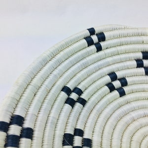 May include: A white woven basket with black stripes. The basket is round and has a wide opening. It is made of natural materials and has a rustic look.