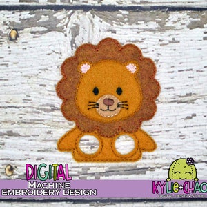 May include: A felt lion finger puppet in shades of brown, tan, and orange. The lion has a detailed face with a tan nose, black eyes, and whiskers. The text "DIGITAL MACHINE EMBROIDERY DESIGN" is at the bottom.
