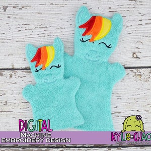 May include: Two light blue terry cloth hand puppets shaped like a pony, with rainbow manes and embroidered facial features. The puppets have a smiling face and are designed for digital machine embroidery. The text "DIGITAL MACHINE EMBROIDERY DESIGN" is visible.